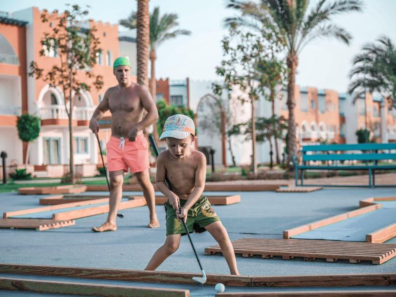 Twee jongens die minigolf spelen op een zonnige buitenbaan bij palmbomen en gebouwen.