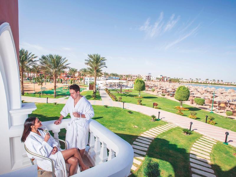 Twee mensen in badjassen op een balkon met uitzicht op een resort met palmbomen en een strand.