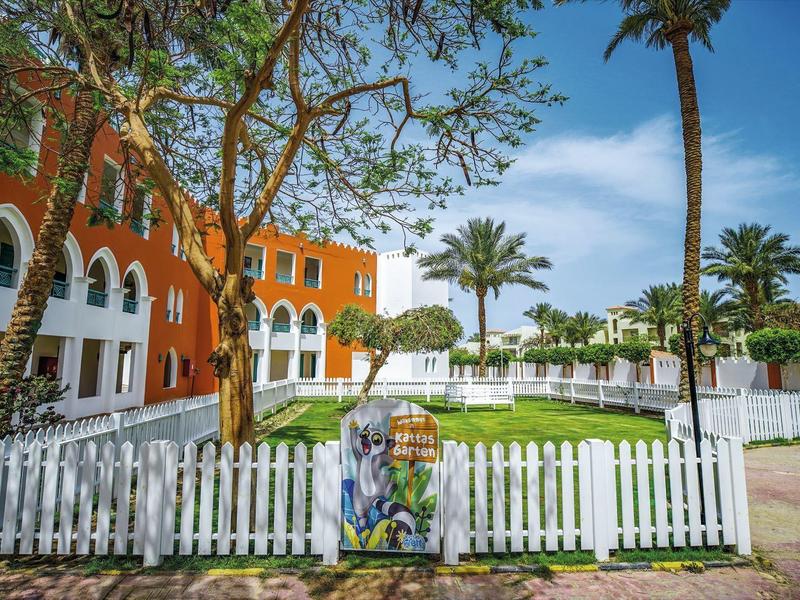 Tuin met witte omheining, palmbomen en oranje gebouw onder blauwe lucht