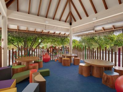 Überdachter Außenbereich mit Kinderspielecken und bunten Möbeln vor grünen Bäumen.