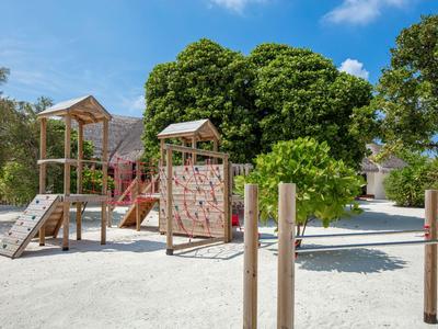 Kinderspielplatz mit Klettergerüst und Rutschen auf weißem Sand, umgeben von Bäumen.
