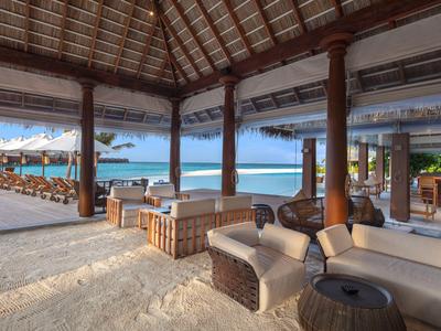 Überdachte Strandlounge mit Sitzbereichen, Blick auf Meer und Sonnenliegen unter blauen Himmel.
