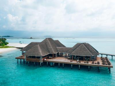 Bungalows auf Stelzen über klarem, blauem Wasser mit einem Steg und Wolkenhimmel.