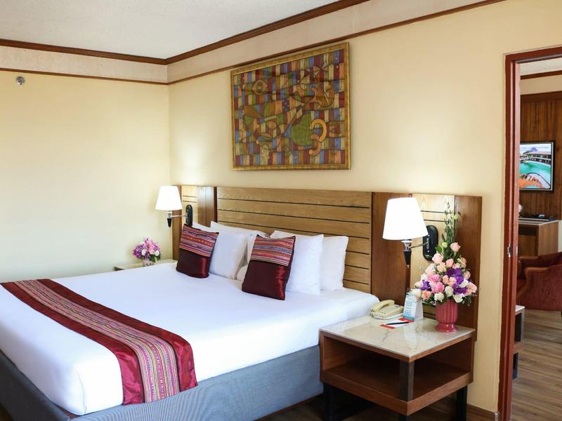 Helles Hotelzimmer mit Doppelbett, Nachttisch, Lampen und Blumenarrangement.