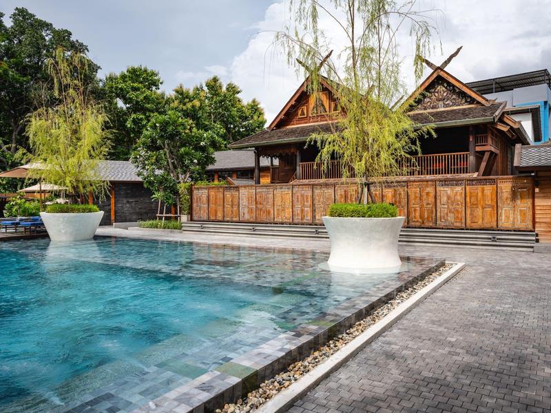 Ein Swimmingpool vor traditionellen Holzhäusern mit Pflanzen in großen weißen Töpfen.