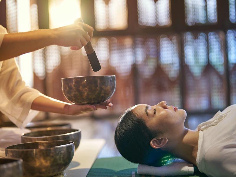 Frau entspannt bei Klangschalentherapie, während Schalen über ihr gehalten werden.