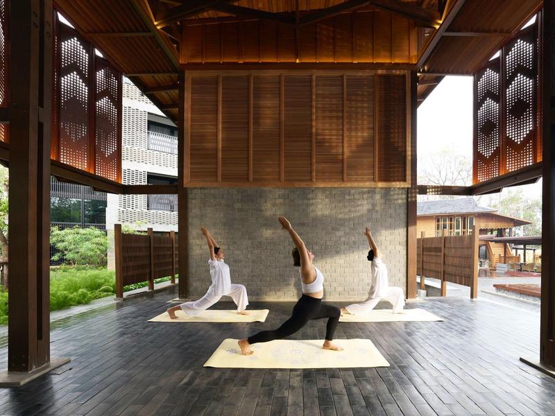 Drei Frauen praktizieren Yoga auf Matten in einer offenen, hölzernen Pavillon-Umgebung.