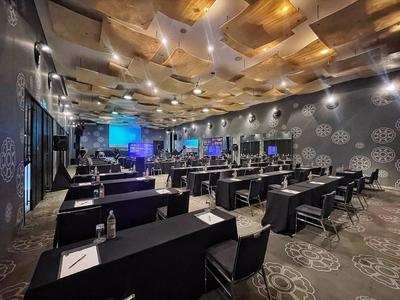 Großer Konferenzraum mit Reihen schwarzer Tische und Stühle, moderner Beleuchtung und Projektoren.