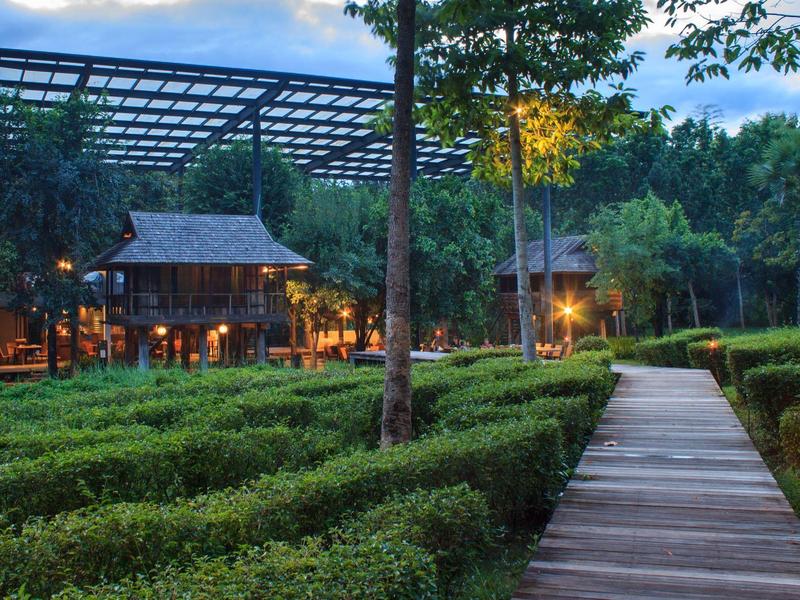 Beleuchteter Holzsteg mit Bäumen und Sträuchern führt zu einem traditionellen Gebäude in der Dämmerung.