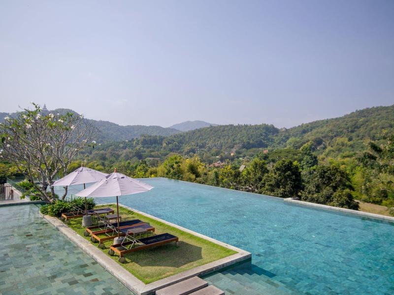 Infinity-Pool mit Liegestühlen und Sonnenschirmen vor bewaldeten Hügeln unter klarem Himmel