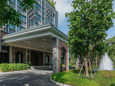 Moderner Gebäudeeingang mit Vordach, grünen Pflanzen, Bäumen und einem Wasserspender im Hintergrund.