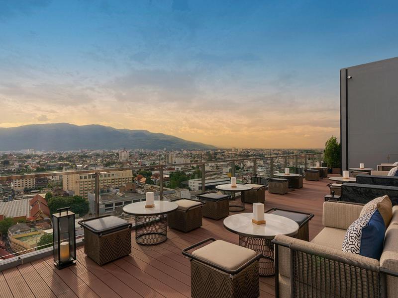 Dachterrasse mit Holzboden, gemütlichen Sitzgruppen und Glasgeländer, Blick auf Stadt und Berge.