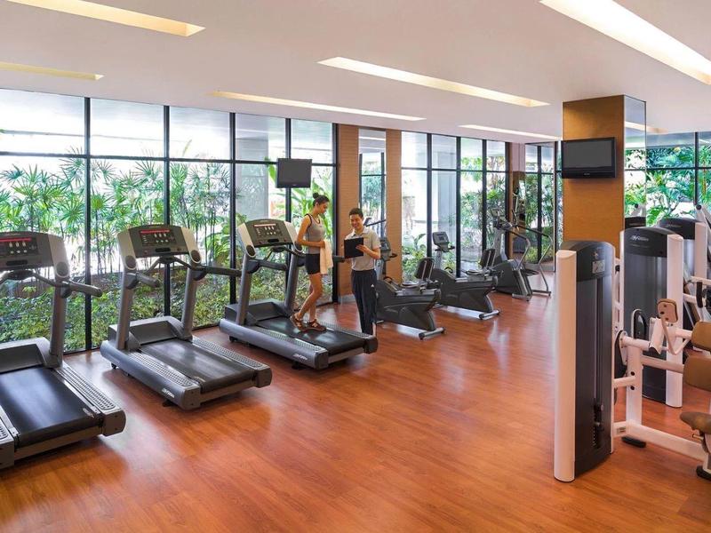 Helles Fitnessstudio mit Laufbändern und Kraftgeräten vor großen Fenstern und Holzboden.