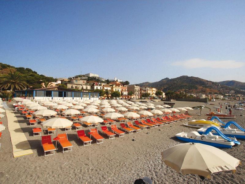 Strand mit Reihen von weißen Sonnenschirmen und orangen Liegen vor einer Küstenstadt bei blauem Himmel.