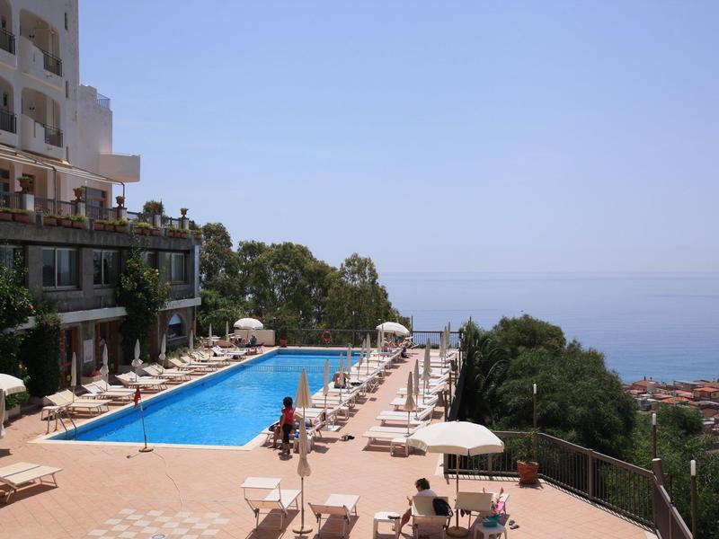 Großer Pool mit Liegestühlen auf Terrasse, links mehrstöckiges Gebäude, weiter Blick auf Meer und Himmel.