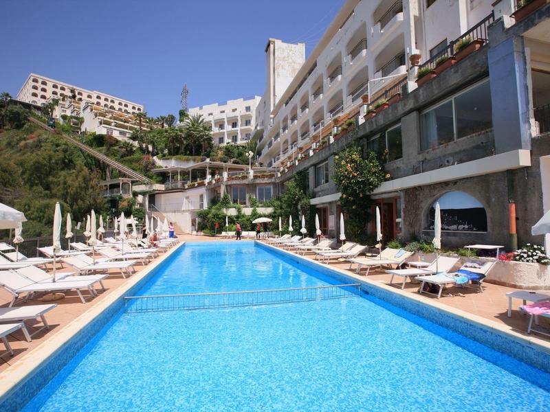 Großer blauer Pool mit weißen Liegestühlen und Sonnenschirmen vor einem mehrstöckigen Hotelgebäude.