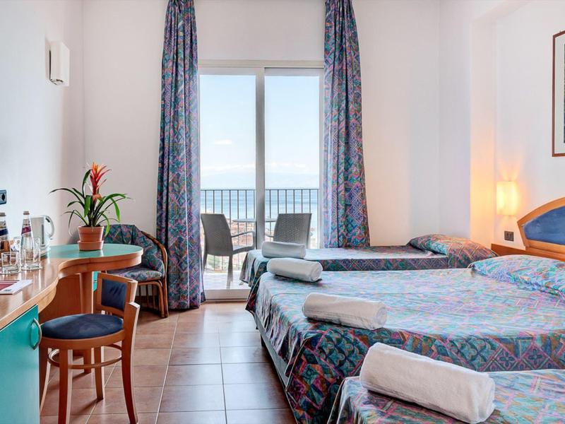 Helles Hotelzimmer mit zwei Einzelbetten, Balkon und Blick aufs Meer, dekoriert mit Vorhängen und Stühlen.