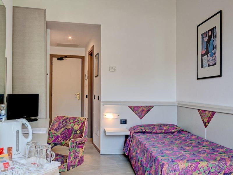 Kleines Zimmer mit Einzelbett, farbig gemusterter Bettdecke und Sessel, kleinem Fernseher und Wandbild.