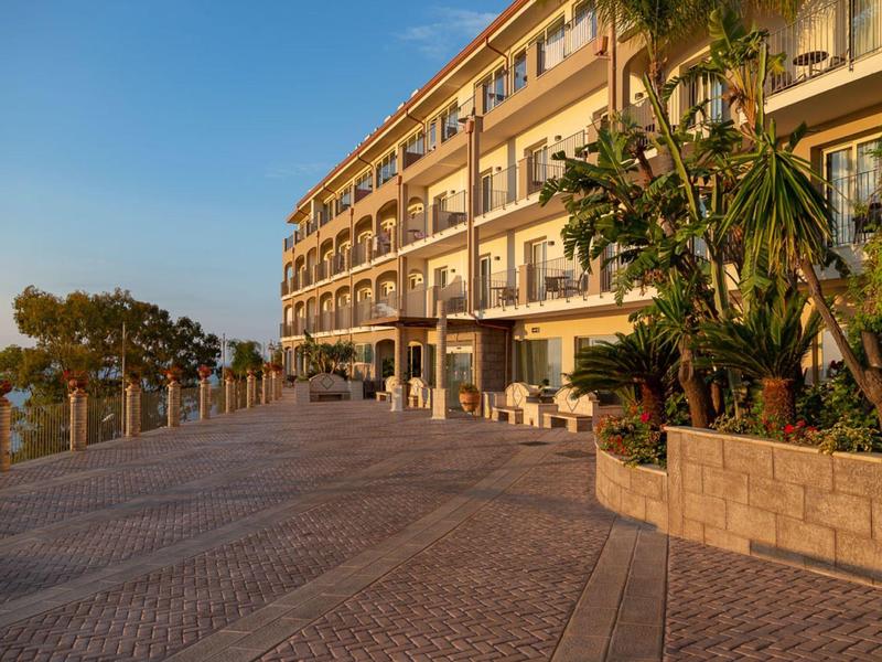 Großes Hotelgebäude am Meer mit sonnigem blauen Himmel und Pflanzkübeln neben breitem Gehweg.