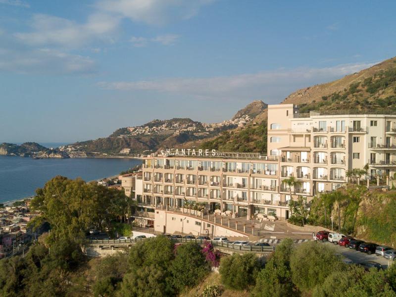 Küstenstadt mit weißen Gebäuden, grünem Hügel und blauem Meer unter hellem Himmel.