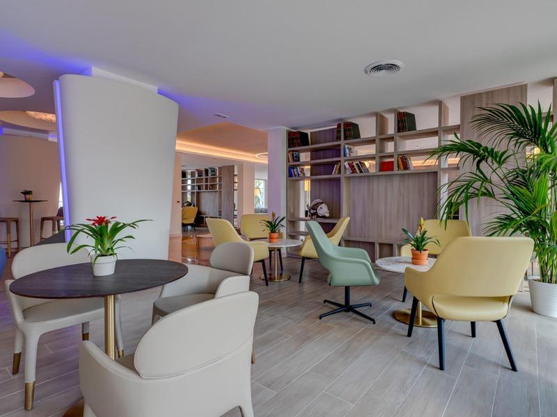 Heller, moderner Raum mit bunten Sesseln, Tischen und grünen Pflanzen, gemütliche Atmosphäre.