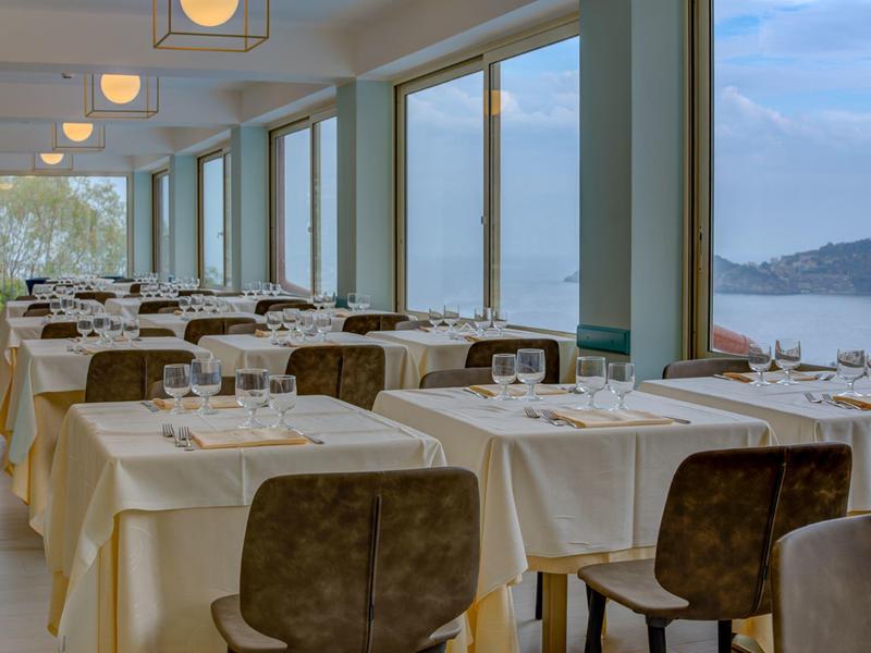 Restaurant mit weißen Tischtüchern, braunen Stühlen und großen Fenstern mit Meerblick.