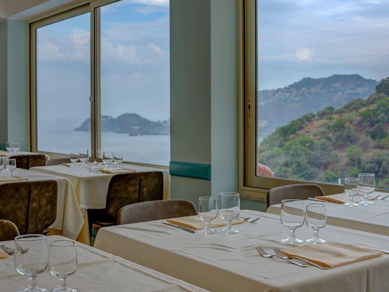 Restaurant mit weißen Tischen und Blick auf Hügel und Meer durch große Fenster.