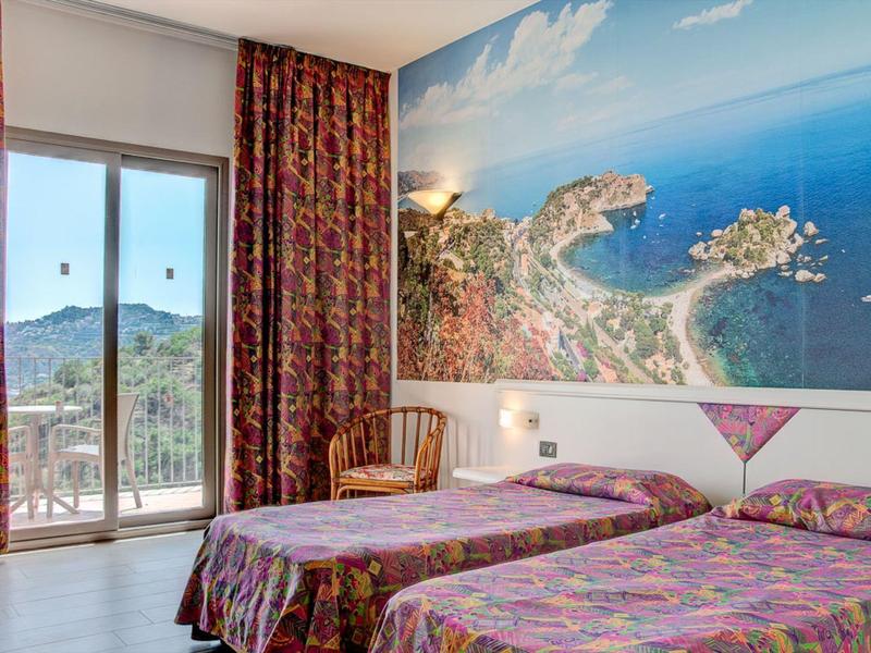 Zimmer mit zwei Einzelbetten, Blumenbettwäsche, großem Fenster und Wandbild einer Küstenlandschaft.