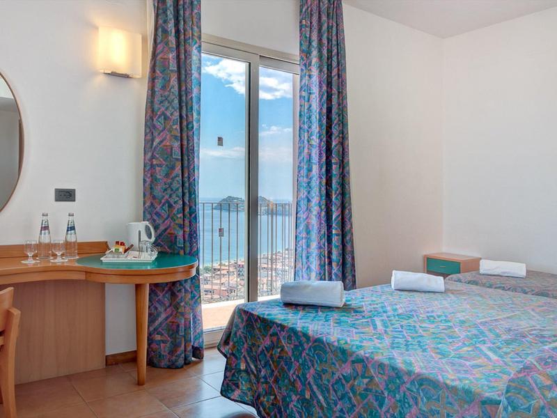Helles Hotelzimmer mit Doppelbett, Schreibtisch, Stuhl, Balkon und Blick auf das Meer bei blauem Himmel.