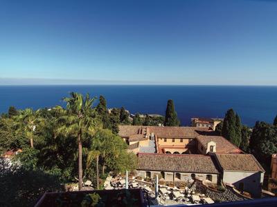 Blick auf mediterranen Küstenort mit Palmen, roten Dächern und tiefblauem Meer unter klarem Himmel.