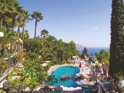 Großer Pool mit blauer Wasserfläche, umgeben von Liegen, weißen Sonnenschirmen, Palmen und Meer im Hintergrund.