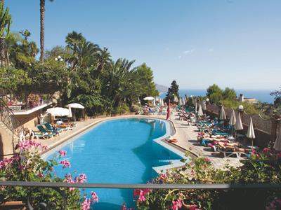 Ein blauer Pool mit Liegestühlen, Sonnenschirmen und Palmen, dahinter Blick aufs Meer.