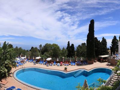 Großes Schwimmbecken mit runden Ecken, blauen Liegestühlen und hohen Bäumen im Hintergrund.