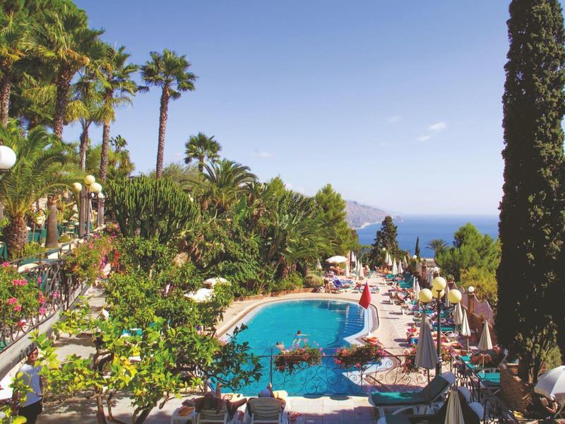 Großer Pool mit blauer Wasserfläche, umgeben von Liegen, weißen Sonnenschirmen, Palmen und Meer im Hintergrund.