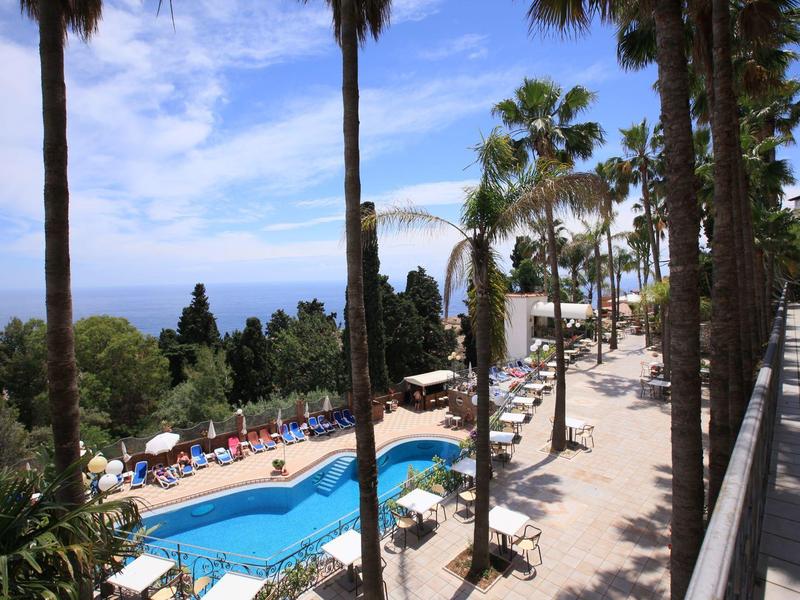 Schwimmbad mit blauer Wasserfläche, umgeben von Palmen und Liegestühlen an einem sonnigen Tag.