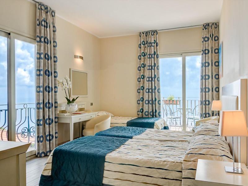 Helles Schlafzimmer mit zwei Betten, blauen Vorhängen und Meerblick durch große Fenster.