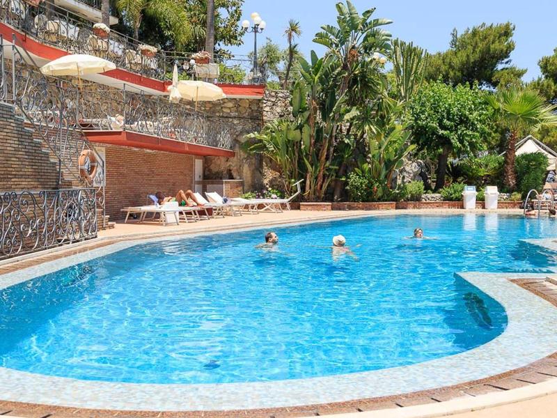 Großer blauer Pool mit Menschen, Liegestühlen und tropischer Bepflanzung im Hintergrund.