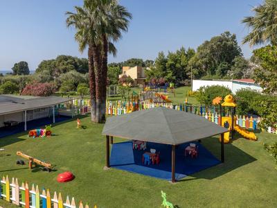 Kinderspielplatz mit Bäumen, Pavillon und buntem Zaun in einem Hotelgarten bei klarem Himmel.
