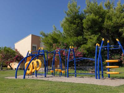 Spielplatz mit blauen und gelben Klettergeräten auf grünem Rasen, umgeben von Bäumen und Gebäuden.