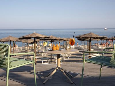 Zwei grüne Stühle und ein Tisch mit Getränken auf einer Terrasse mit Blick auf einen Sandstrand und das Meer.