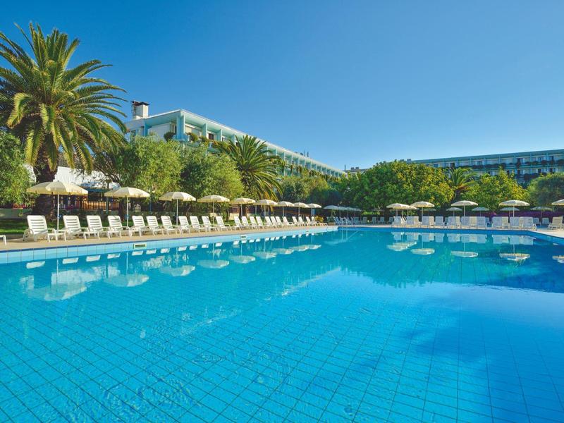Groot buitenzwembad met ligstoelen en parasols naast een hotel onder een heldere blauwe lucht.