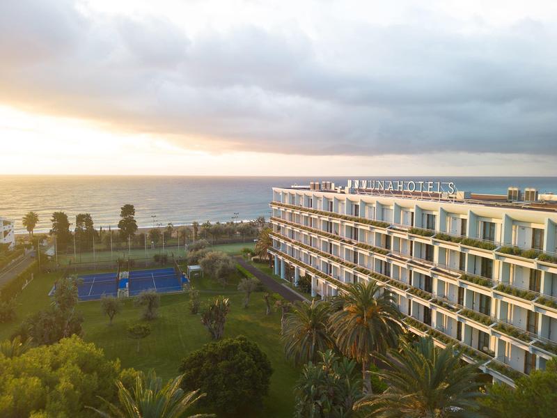 Hotel aan zee met een tennisbaan, palmbomen en een bewolkte lucht bij zonsondergang.