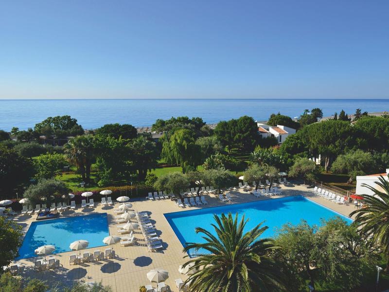 Groot buitenzwembad met ligstoelen en zeezicht, omgeven door palmbomen en groen.