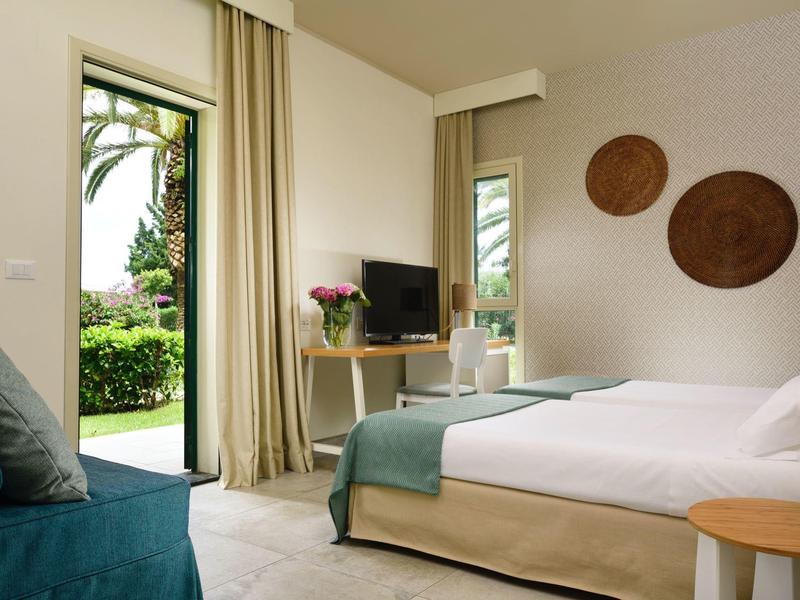 Lichte hotelkamer met tweepersoonsbed, groot raam, tv en uitzicht op tuin.