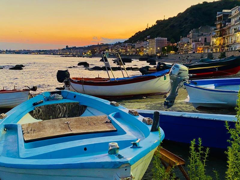 Kleurrijke boten aangemeerd langs de kustlijn bij zonsondergang met gebouwen en heuvels op de achtergrond.