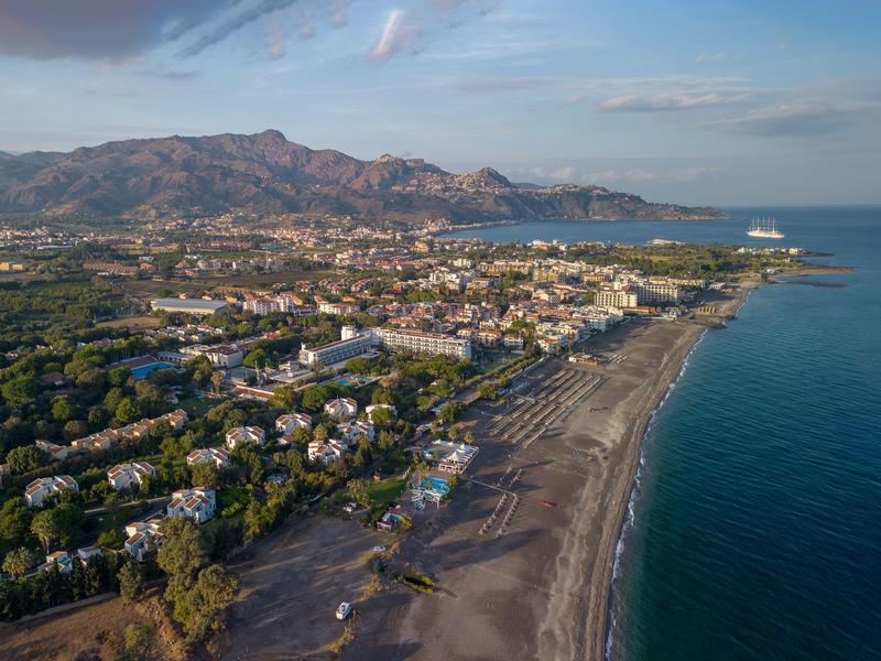 Een kustplaats met een zandstrand, blauwe zee en bergen op de achtergrond onder een gedeeltelijk bewolkte lucht.