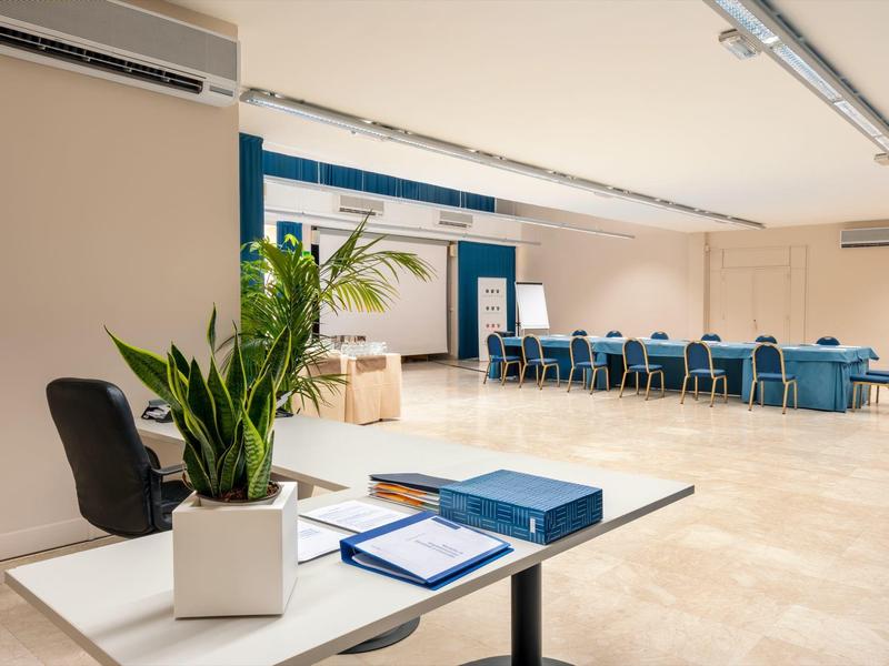 Ruime, goed verlichte vergaderruimte met tafels, stoelen en een potplant op de voorgrond.