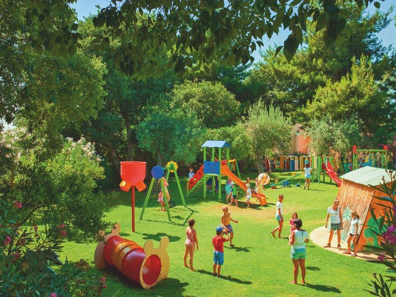 Kinderen spelen in een groene buitenspeelplaats omgeven door bomen en struiken.