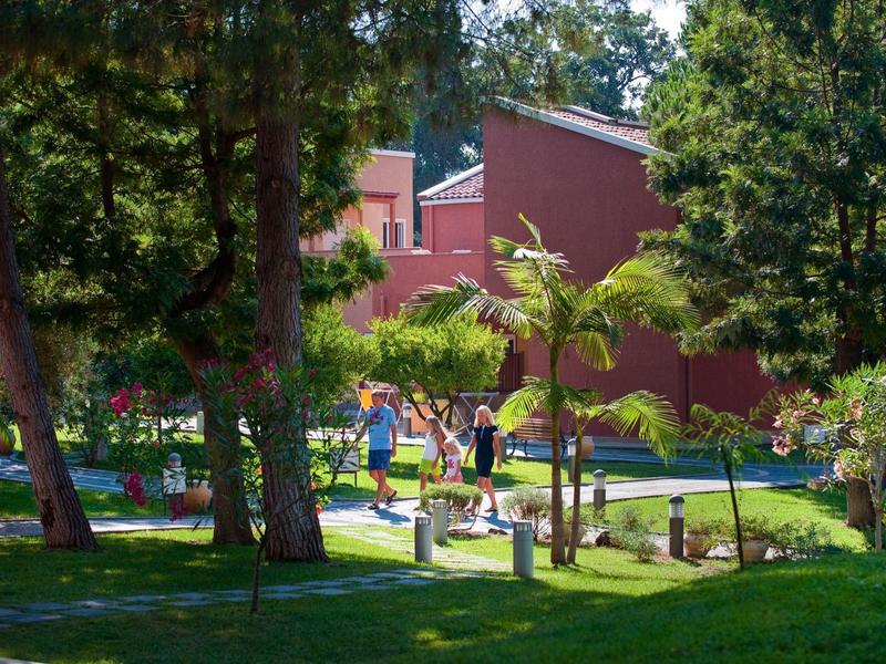 Mensen wandelen in een zonnige tuin met bomen en rode gebouwen in de buurt.