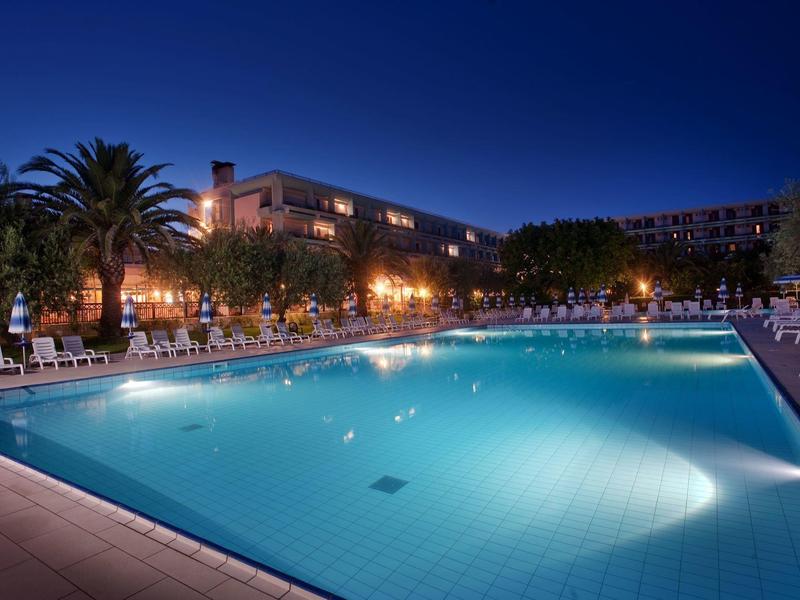 Groot buitenzwembad 's nachts verlicht met omliggende palmbomen en hotelgebouw.